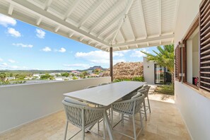 En overdækket terrasse med borde og stole, udsigt over landskabet og en bygning med stråtækt tag.