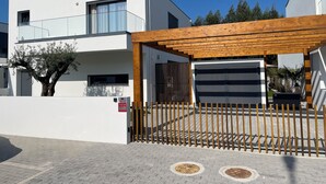 Un bâtiment moderne à deux étages, doté d’une façade blanche, d’une pergola en bois au-dessus de l’entrée et d’une allée d’accès fermée par un portail.