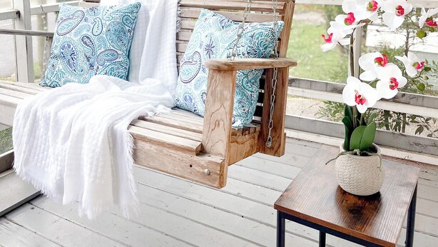 A wooden swing chair with patterned cushions, a white throw blanket, and a small table with a vase of white flowers on a porch. - Kalmia Suite A - Centrally Located Downtown (Laurel)