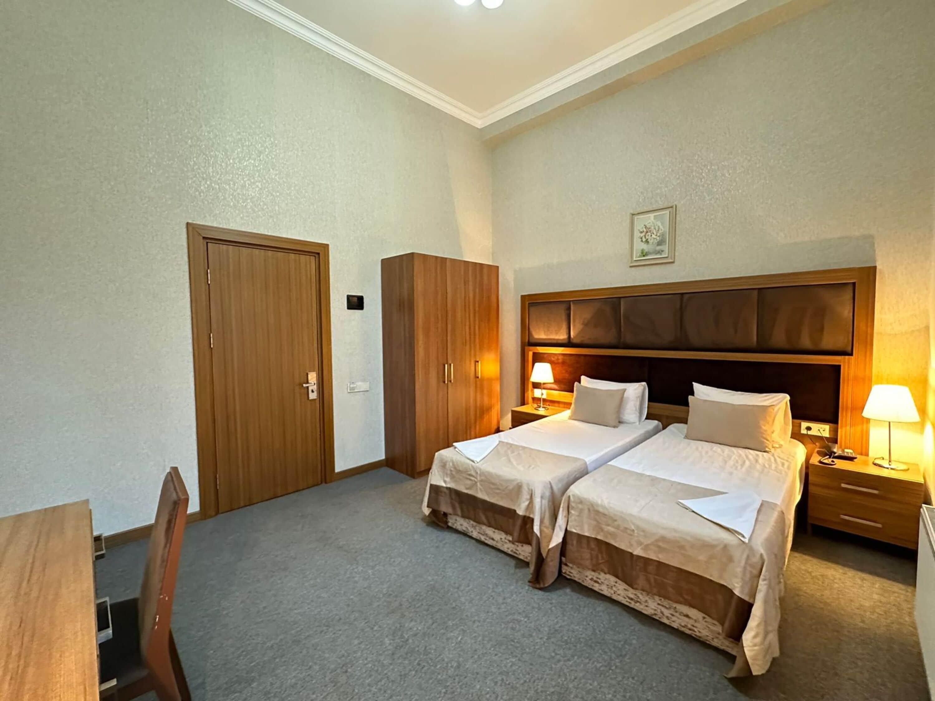 Hotellihuone, jossa on kaksi erillistä sänkyä, puinen vaatekaappi, työpöytä tuolilla sekä yöpöytä lampulla.