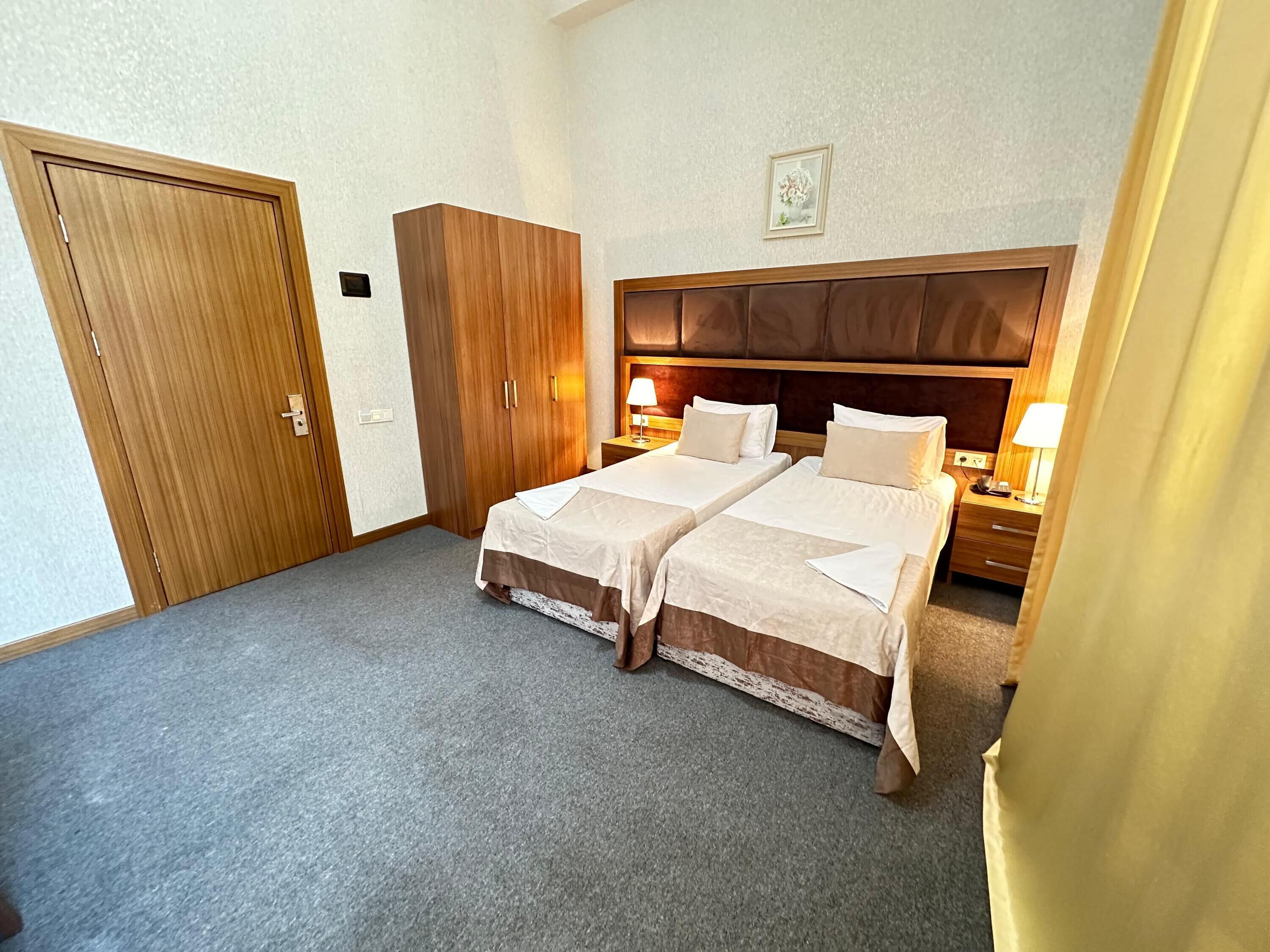 A hotel room with two beds, a wooden wardrobe, a framed picture on the wall, and a nightstand with a lamp.