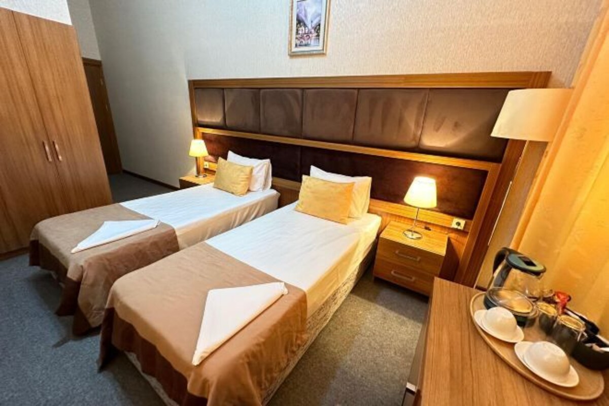 Una habitación de hotel con dos camas individuales, un armario de madera, una mesita de noche con lámpara y una bandeja con platos y una taza.