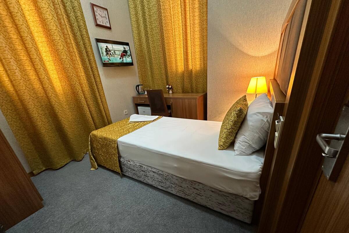 A hotel room with a bed, a desk, a chair, a lamp, curtains, and a clock on the wall.