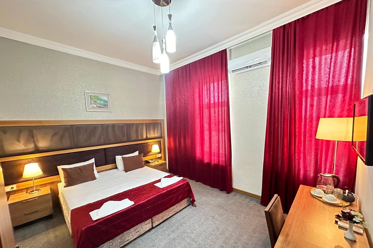 A hotel room with a bed, bedside lamps, a desk with a lamp, and a window with red curtains.