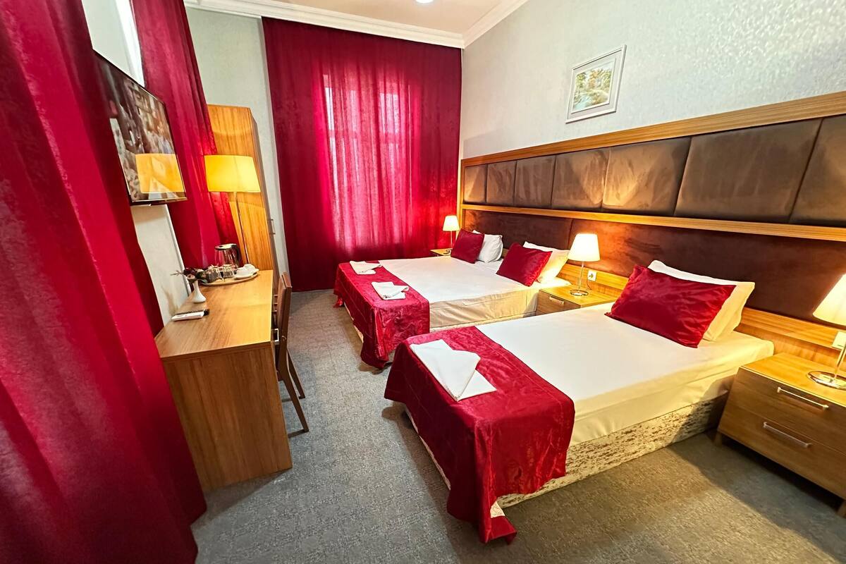 A hotel room with two beds, each with red bedspreads and white linens, a wooden desk with a lamp, and a large window with red curtains.
