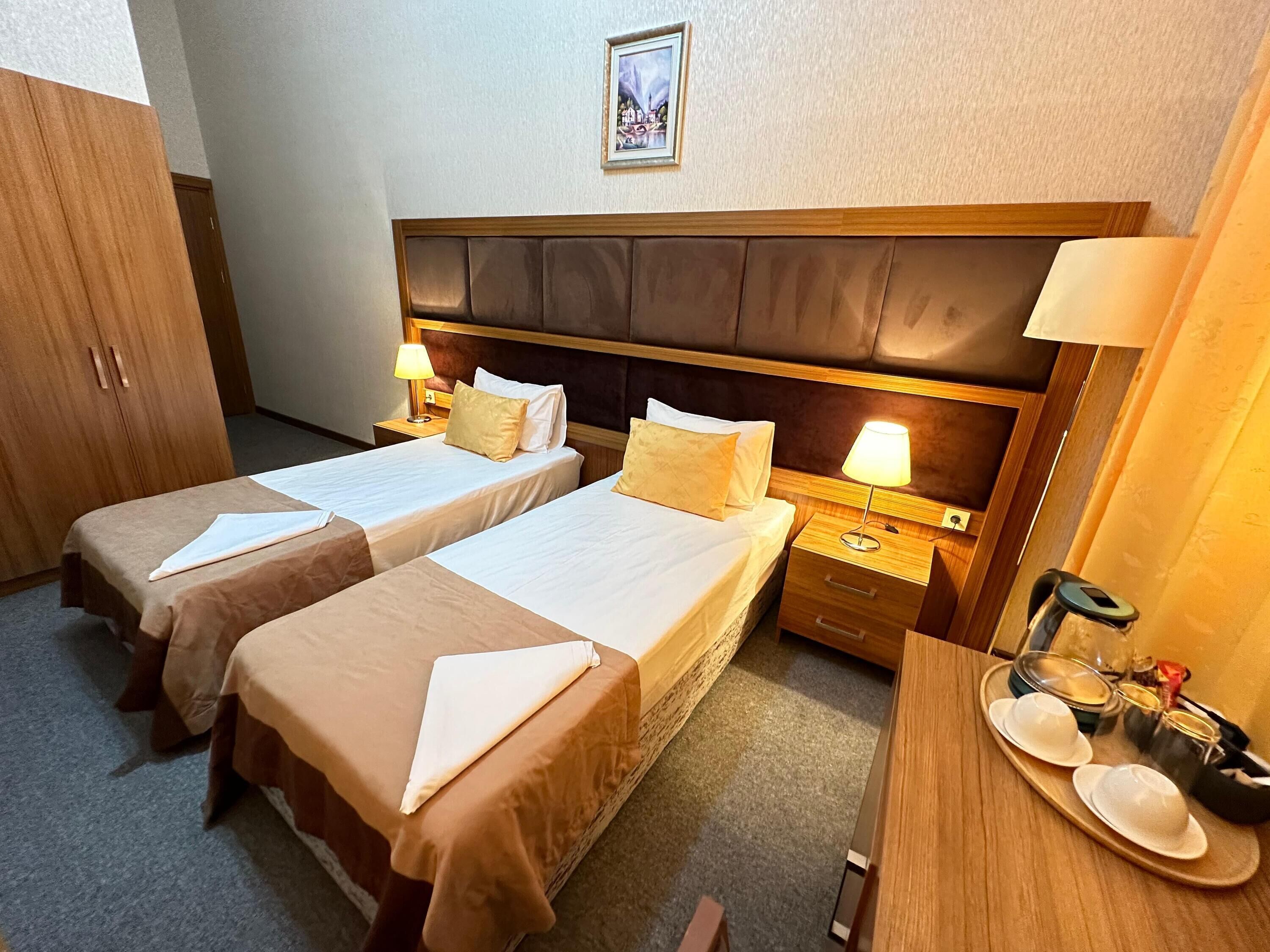 A hotel room with two single beds, a wooden headboard, bedside tables with lamps, a wardrobe, and a tray with cups and a teapot.