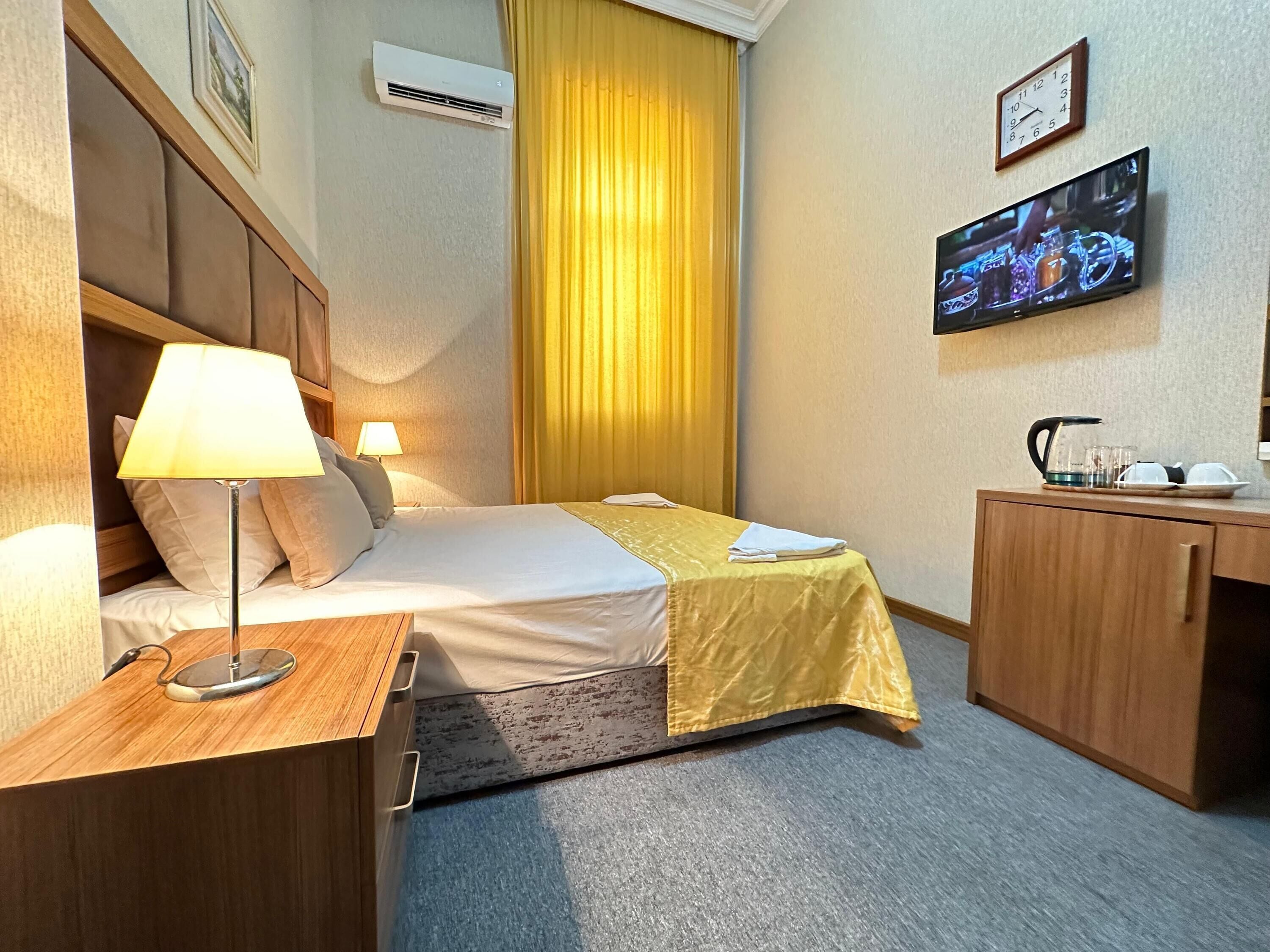 Habitación de hotel con cama, mesita de noche, lámpara, televisor y un escritorio pequeño con hervidor y tazas.