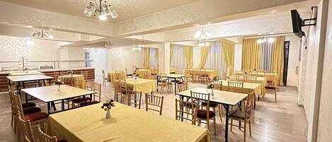 A spacious dining hall with multiple tables and chairs, a chandelier, and curtains on the windows.