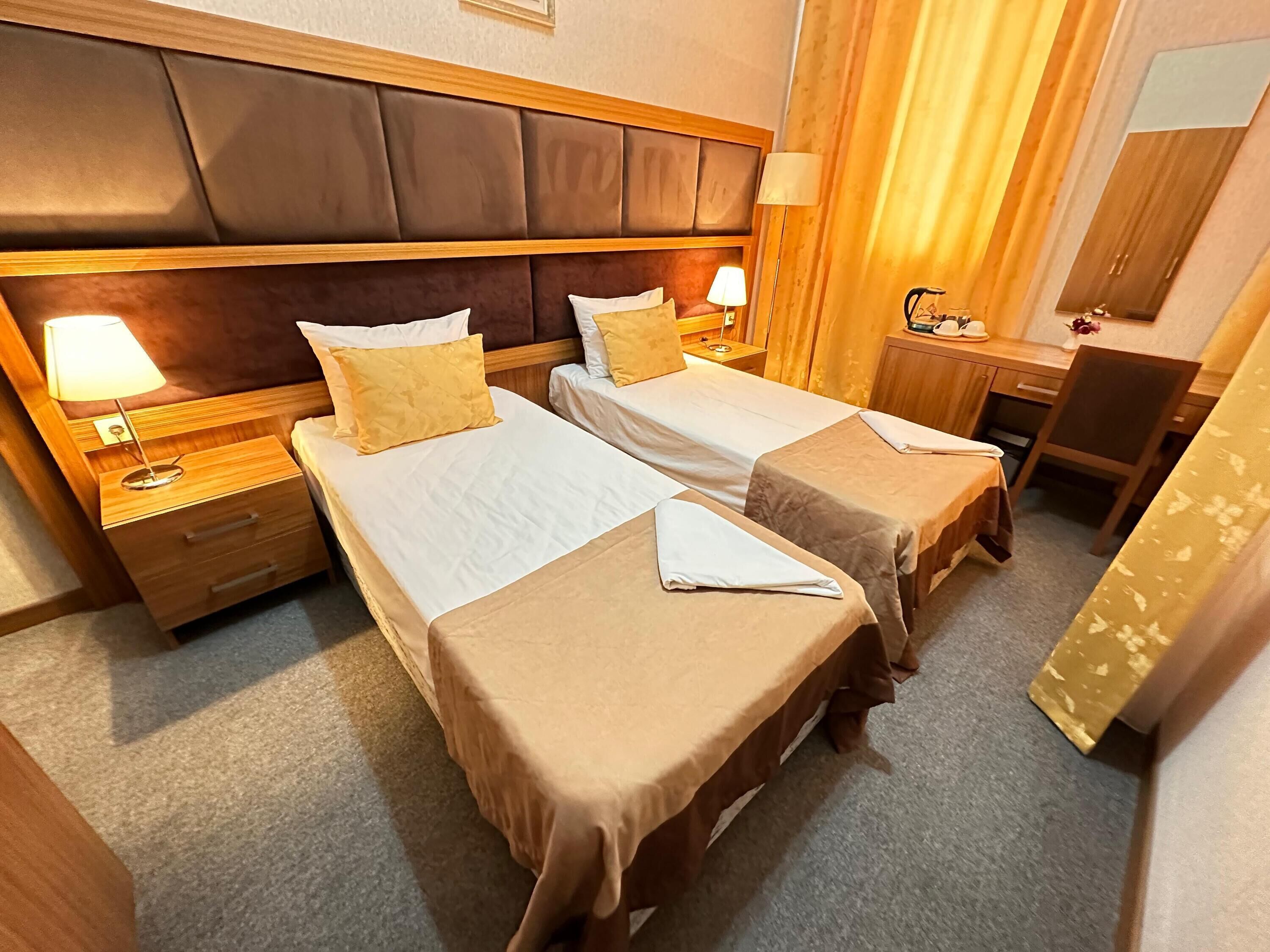 A hotel room with two beds, wooden headboards, bedside tables with lamps, a desk, and a chair.