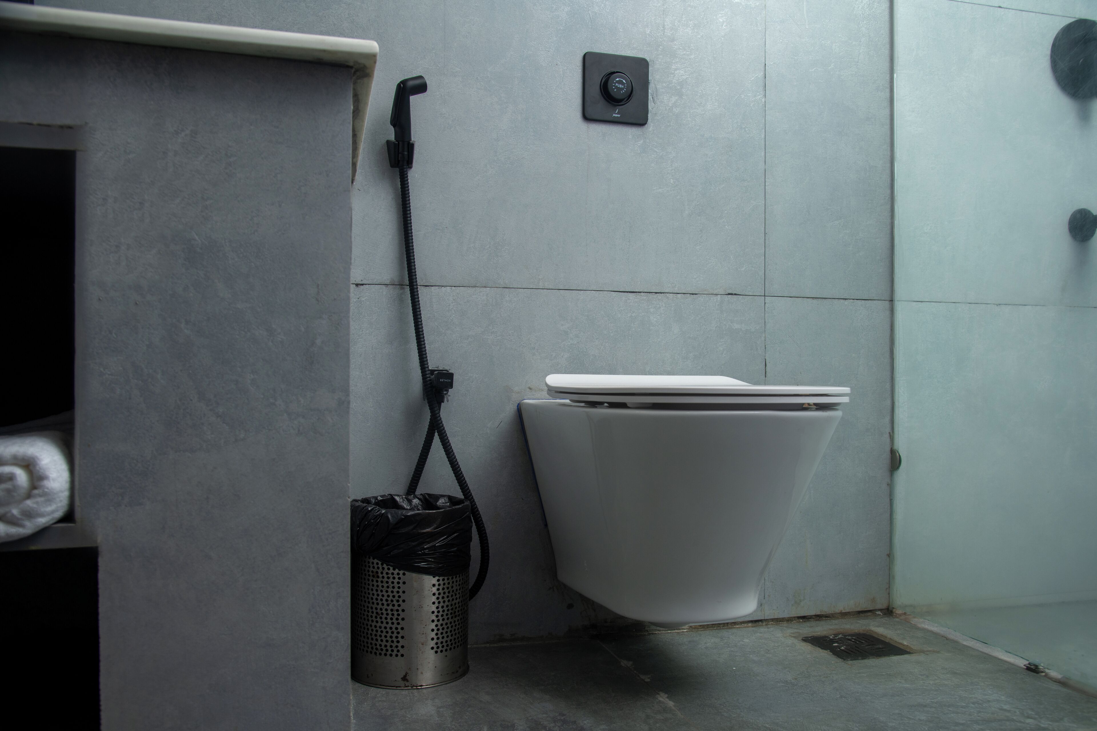Un baño con inodoro, un cubo de basura y un botón de descarga en la pared.