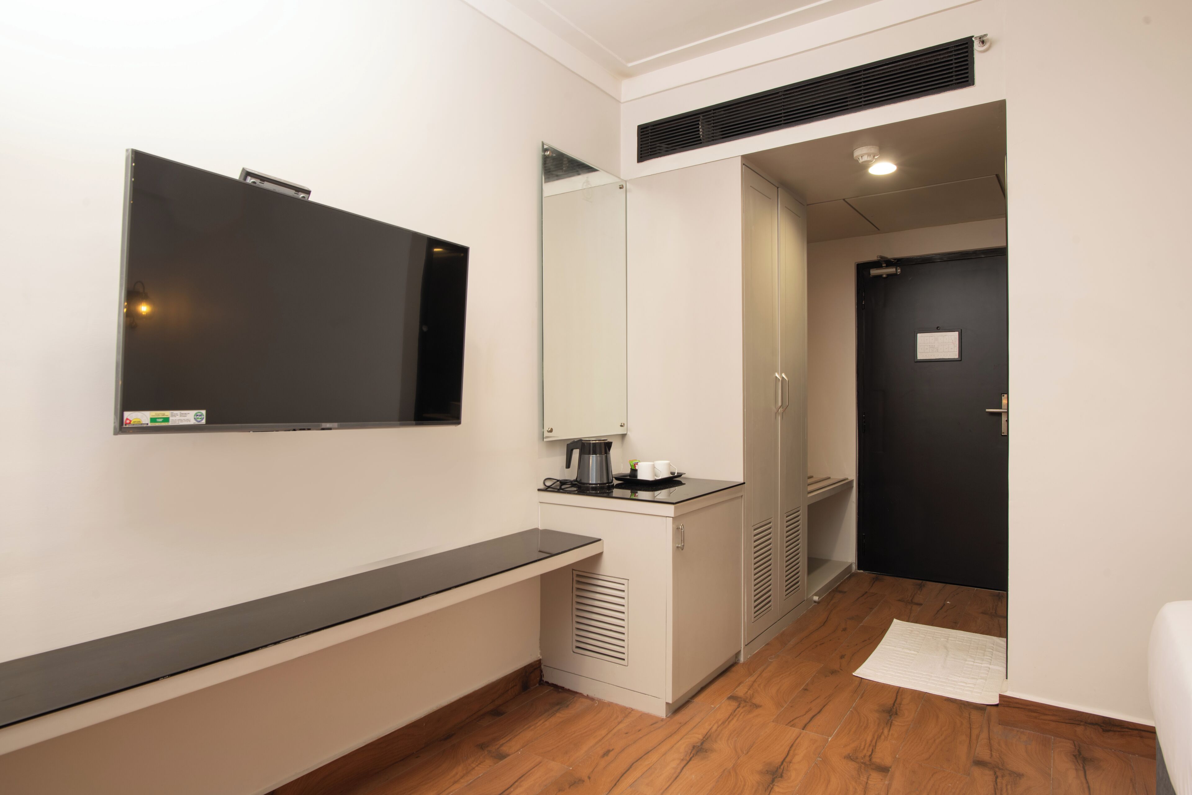 A modern hotel room with a flat-screen TV mounted on the wall, a small kitchenette with a coffee maker, and a door leading to another room.