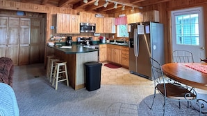 A wooden cabin interior with a kitchen, dining area, and a large window.