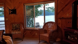 A cozy cabin interior with a large window overlooking a lake, wicker furniture, and a fireplace.