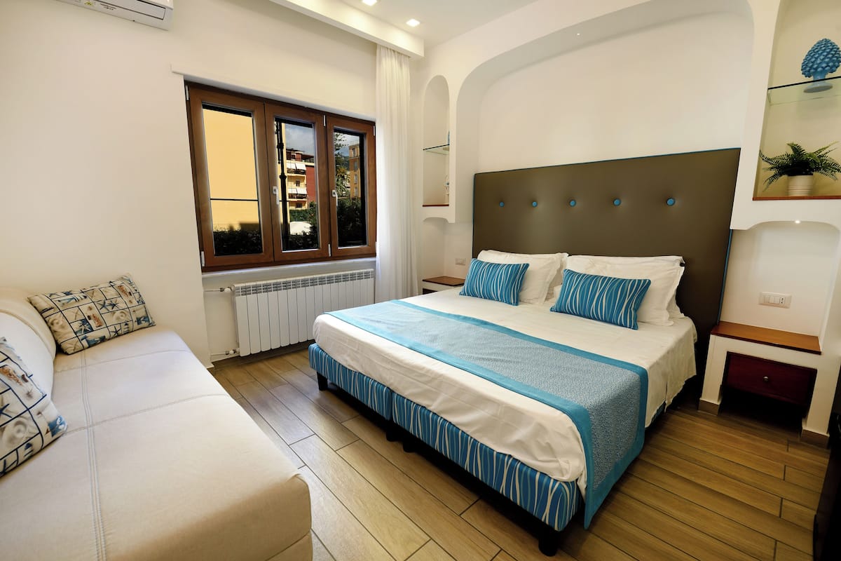 Un dormitorio con una cama grande, una mesita de noche, una ventana con cortinas, un radiador y un sofá.