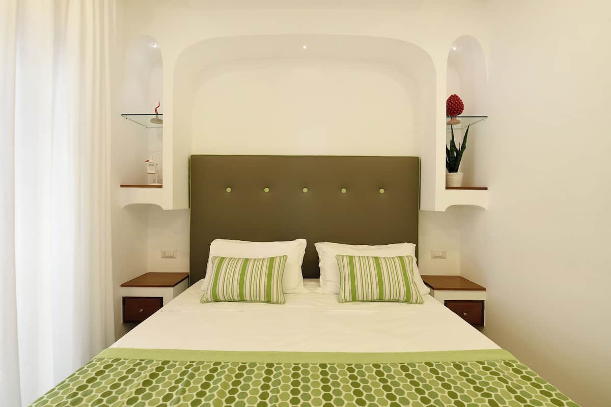 Una cama bien hecha con dos almohadas, una colcha verde estampada y un cabecero marrón. Hay dos mesitas de noche con lámparas y un pequeño estante con una planta.