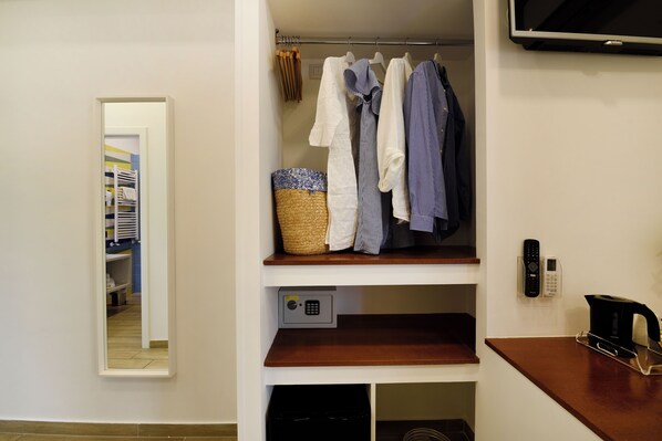 A hotel room with a wardrobe, a mirror, a shelf with clothes, a basket, a phone, and a coffee maker.