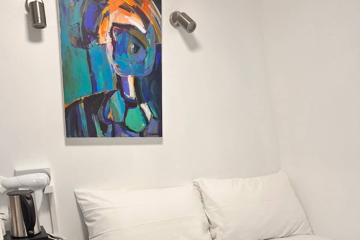 A hotel room with a bed, white bedding, a painting on the wall, a hairdryer, and a coffee maker.