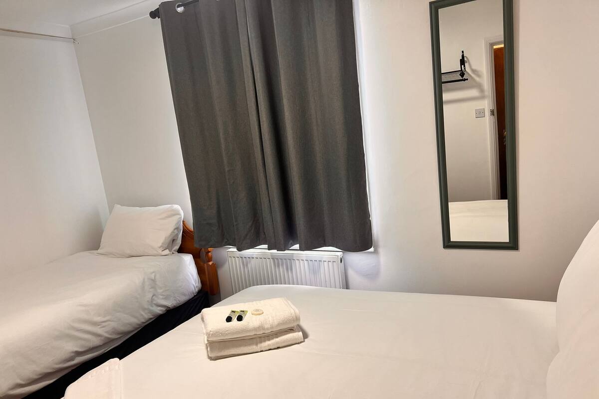 A hotel room with two single beds, a large window with grey curtains, a white radiator, and a mirror on the wall.