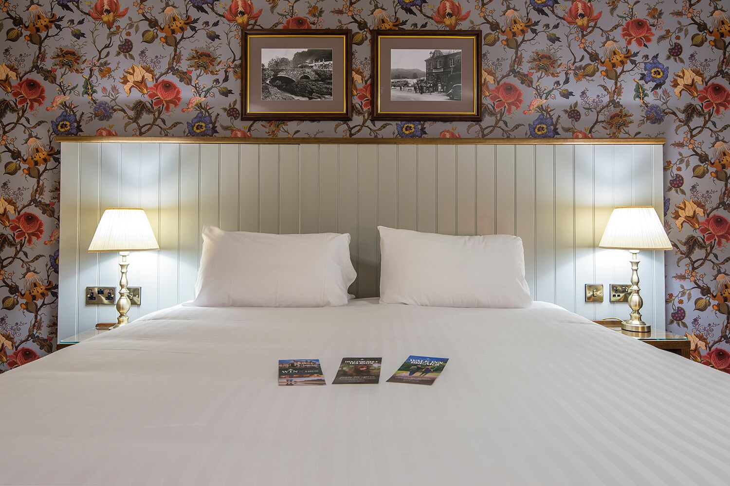 A hotel room with a large bed, two white pillows, two bedside lamps, and floral wallpaper.