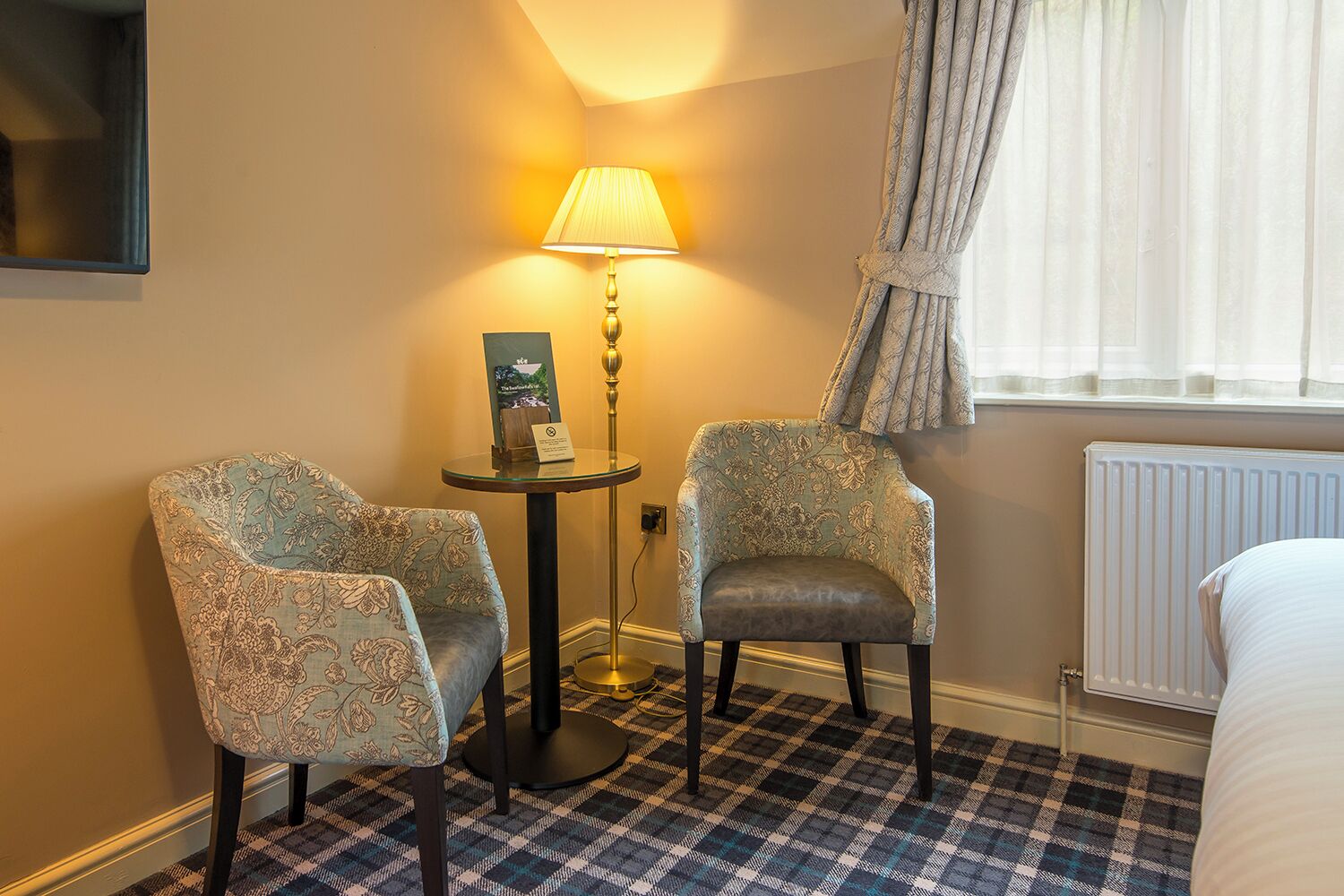 Une chambre d’hôtel avec deux fauteuils rembourrés, une petite table ronde avec une lampe, un tableau encadré et un lit avec une couette blanche.