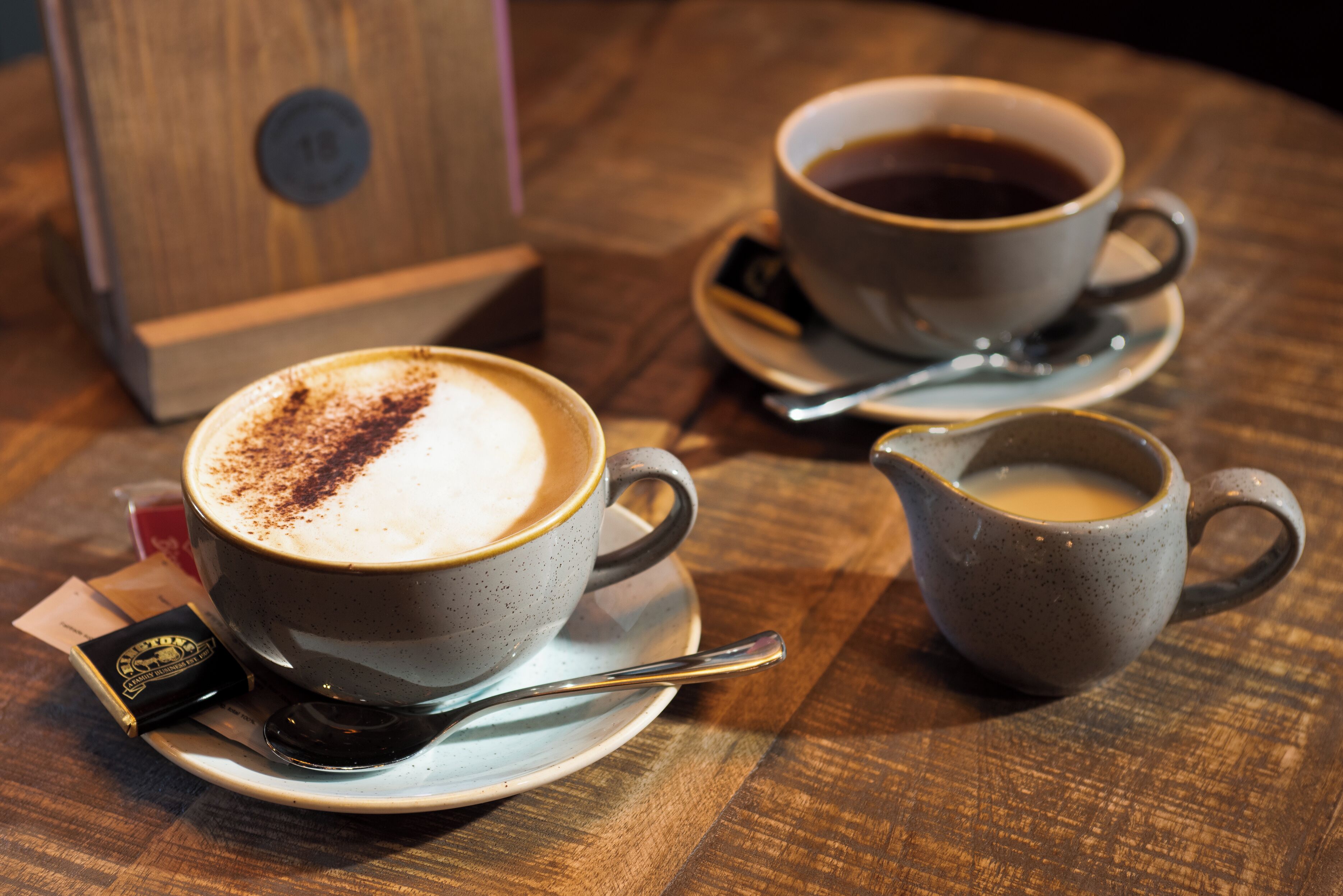 Two cups of coffee on saucers with spoons, a small pitcher, and a wooden box on a wooden table.
