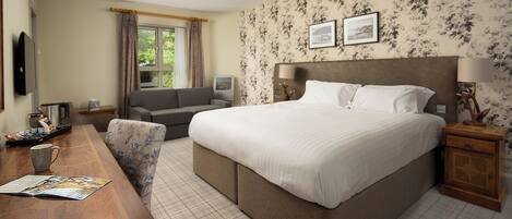 A hotel room with a large bed, a sofa, a desk with a cup and magazine, and a TV.