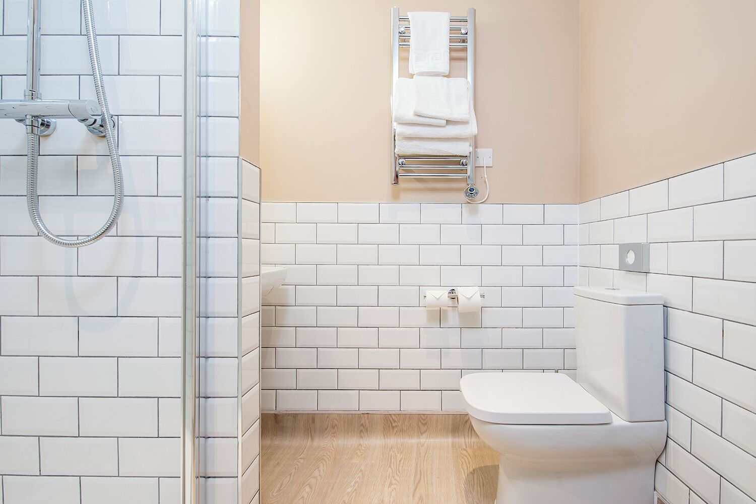 A bathroom with white tiled walls, a wooden floor, a white toilet, a towel rack with towels, and a shower with a hose.