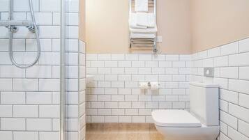 Un baño con paredes de azulejos blancos, suelo de madera, inodoro blanco, toallero con toallas y ducha con manguera.