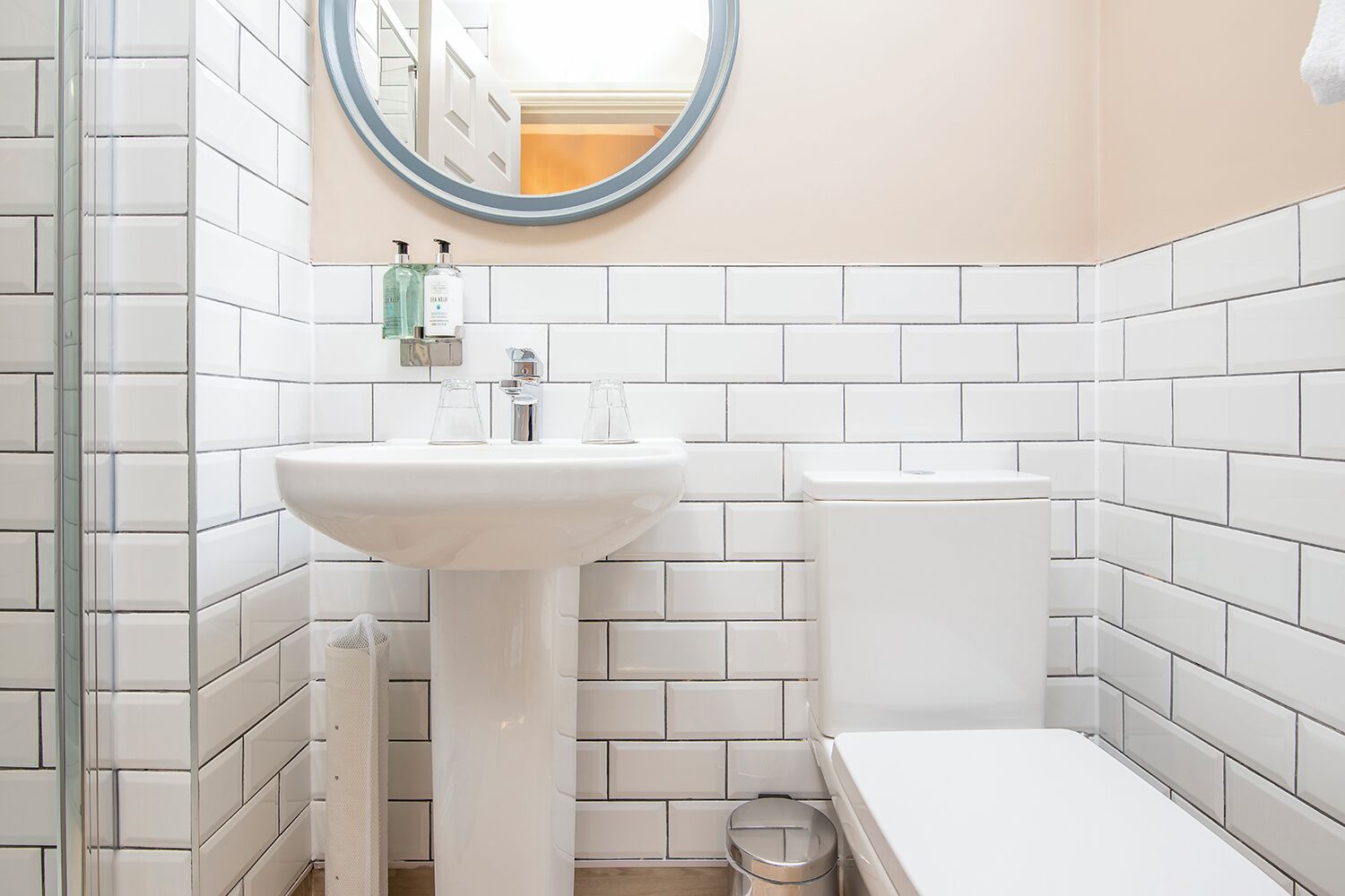 Ein Badezimmer mit weißen Fliesen, einem Standwaschbecken, einer Toilette und einem Mülleimer.