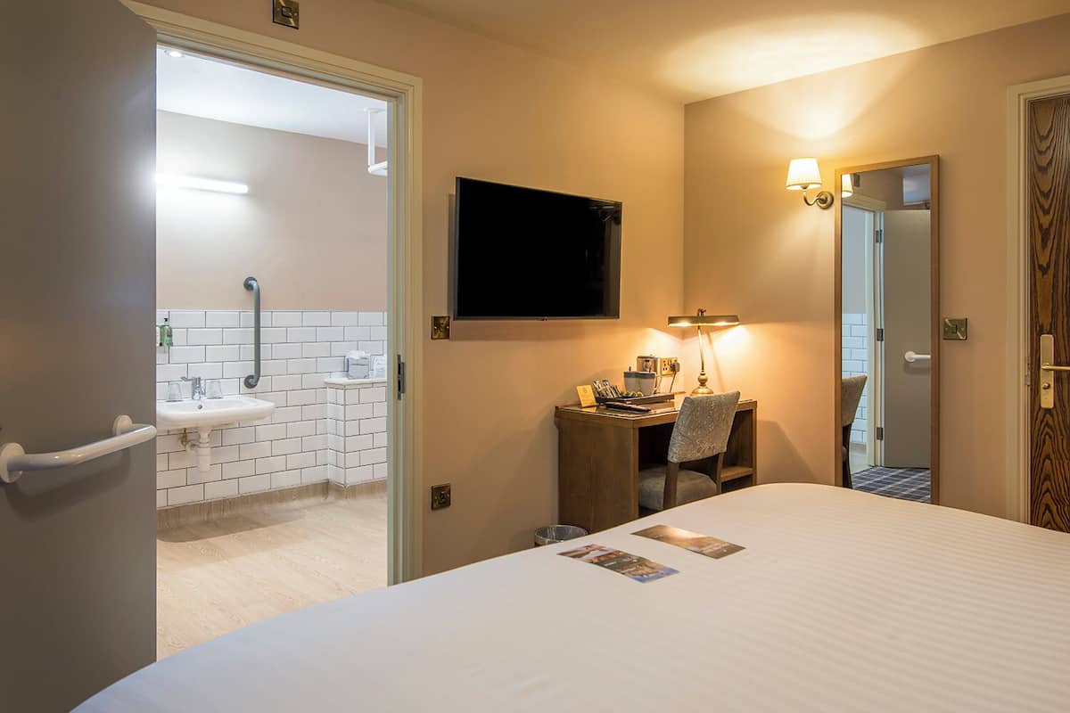 A hotel room with a bed, a desk with a chair, a television mounted on the wall, a mirror, and a bathroom with a sink and toilet.