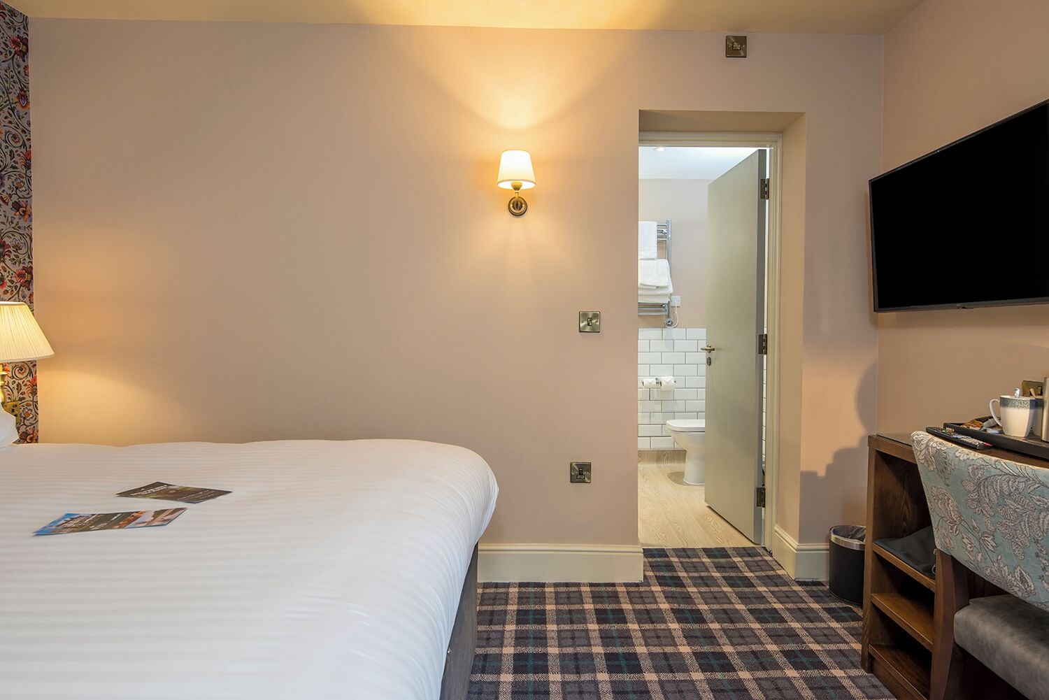 Une chambre d’hôtel avec un lit, un téléviseur fixé au mur, un bureau avec une chaise et une salle de bain visible à travers une porte ouverte.