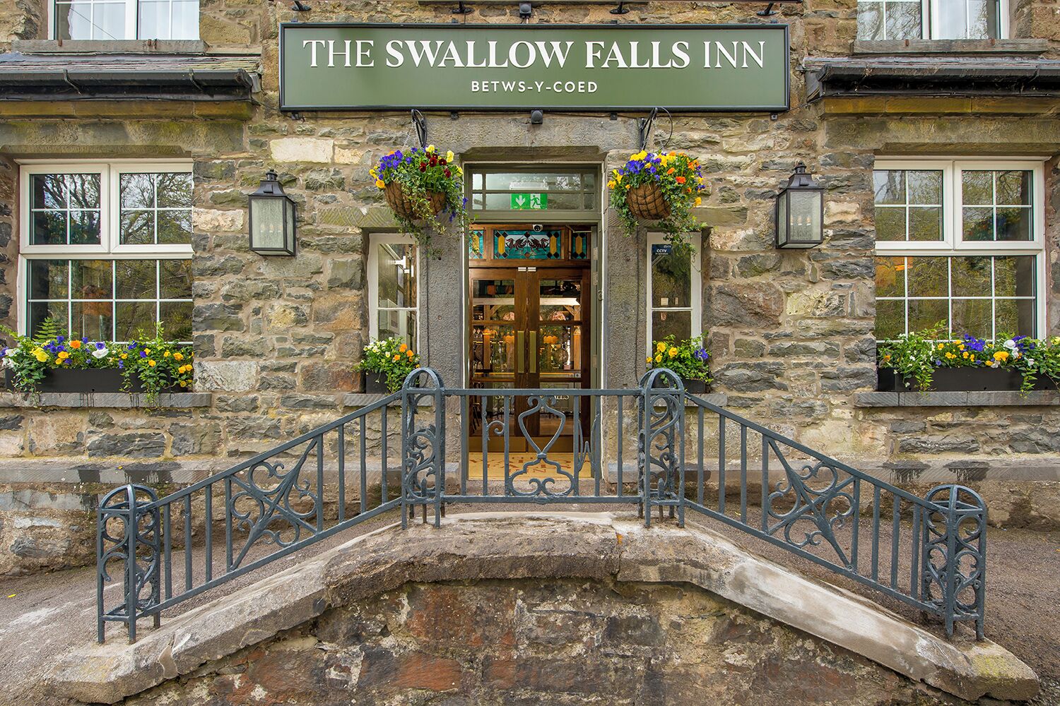 The Swallow Falls Inn, a stone building with a sign above the entrance, a small bridge, and flower baskets on the windowsills.