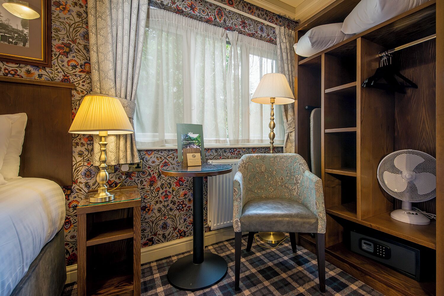 A hotel room with a bed, two lamps, a chair, a small table, a radiator, a fan, and a wardrobe.