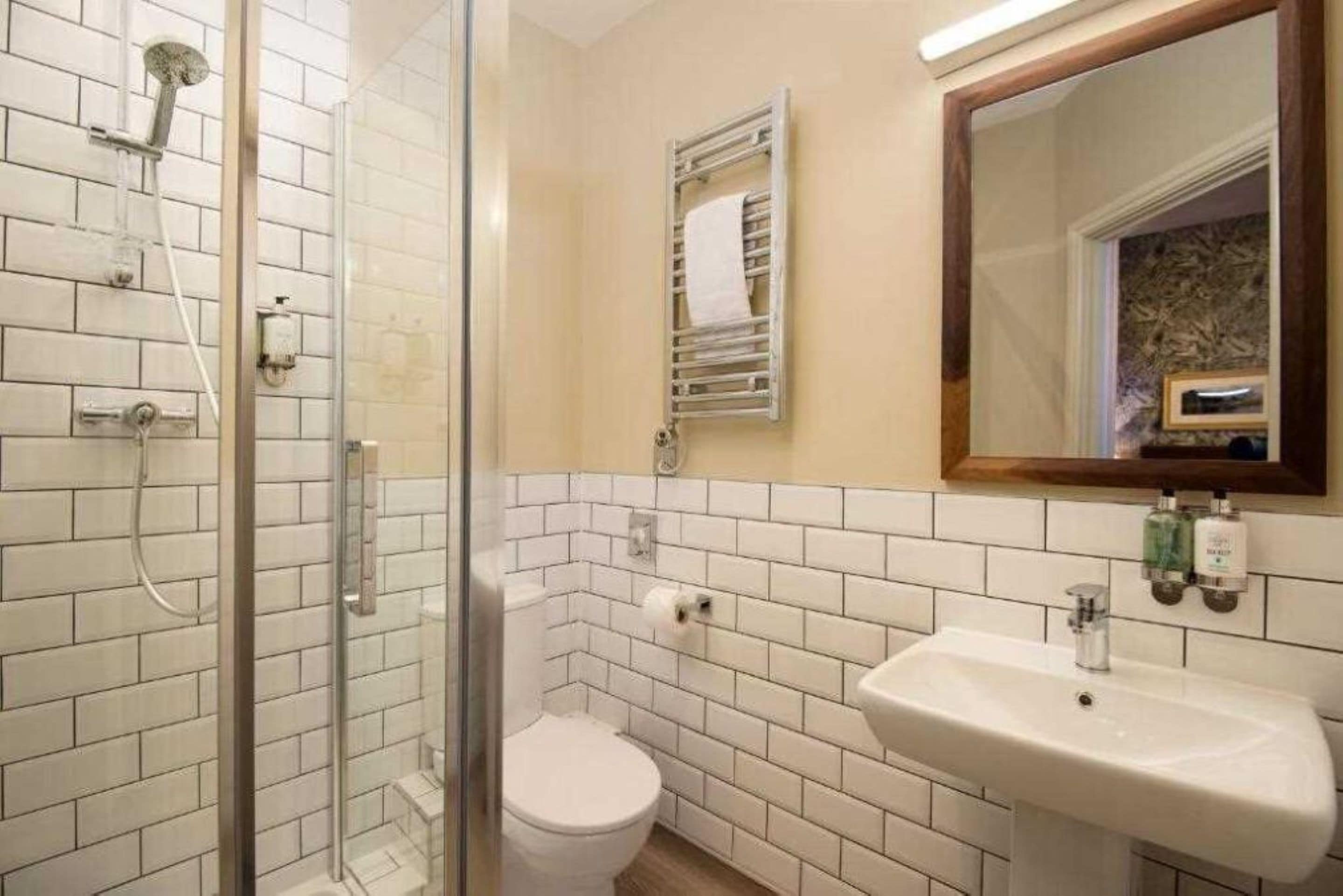 Ein Badezimmer mit einer gläsernen Duschkabine, weißen Fliesenwänden, einer weißen Toilette und einem Waschbecken mit Spiegel darüber.