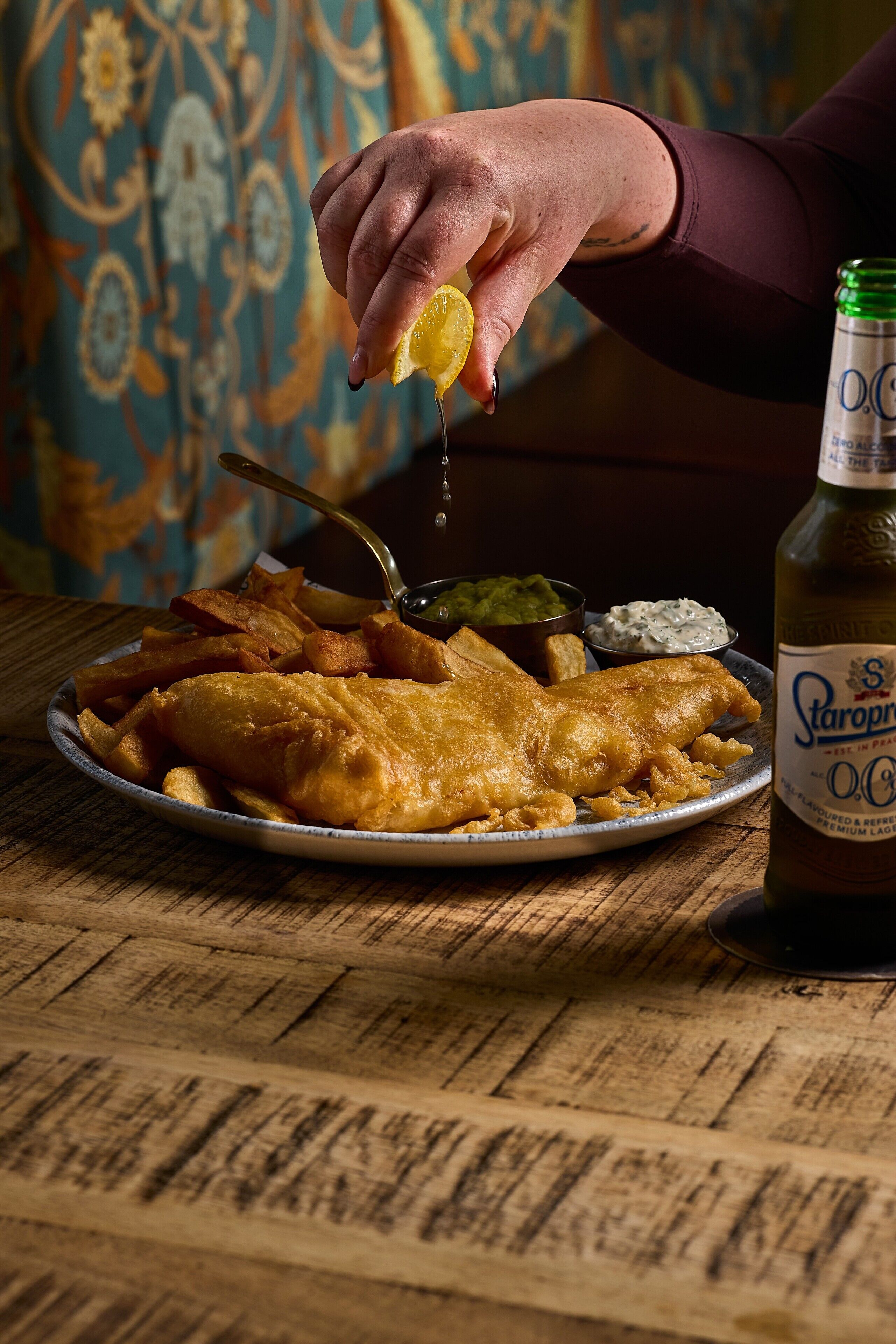 A plate of fish and chips with a bottle of beer and a hand squeezing a lemon.