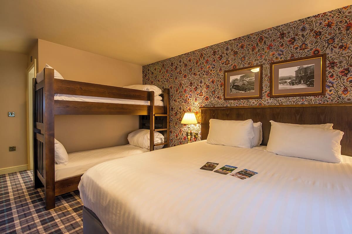 A hotel room with a bunk bed, a large bed with white linens, a patterned wallpaper, and two framed pictures on the wall.