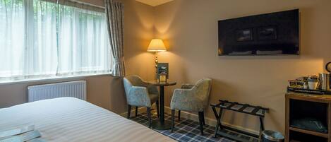 A hotel room with a bed, two chairs, a table, a lamp, a painting, a radiator, a window with curtains, and a check-in counter.