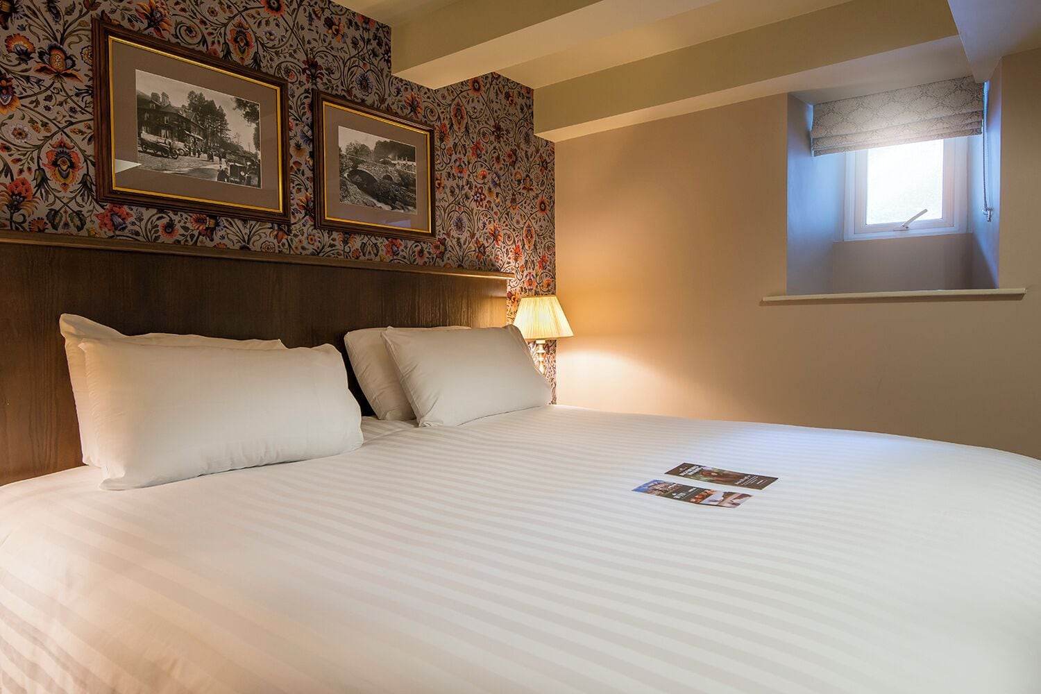 Un lit bien fait, avec du linge de lit blanc, une tête de lit ornée d’un motif floral et deux photographies encadrées accrochées au mur.