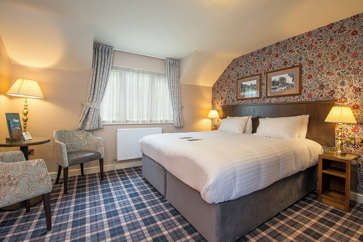 A hotel room with a large bed, two bedside tables with lamps, a chair, a window with curtains, and a floral wallpaper design.