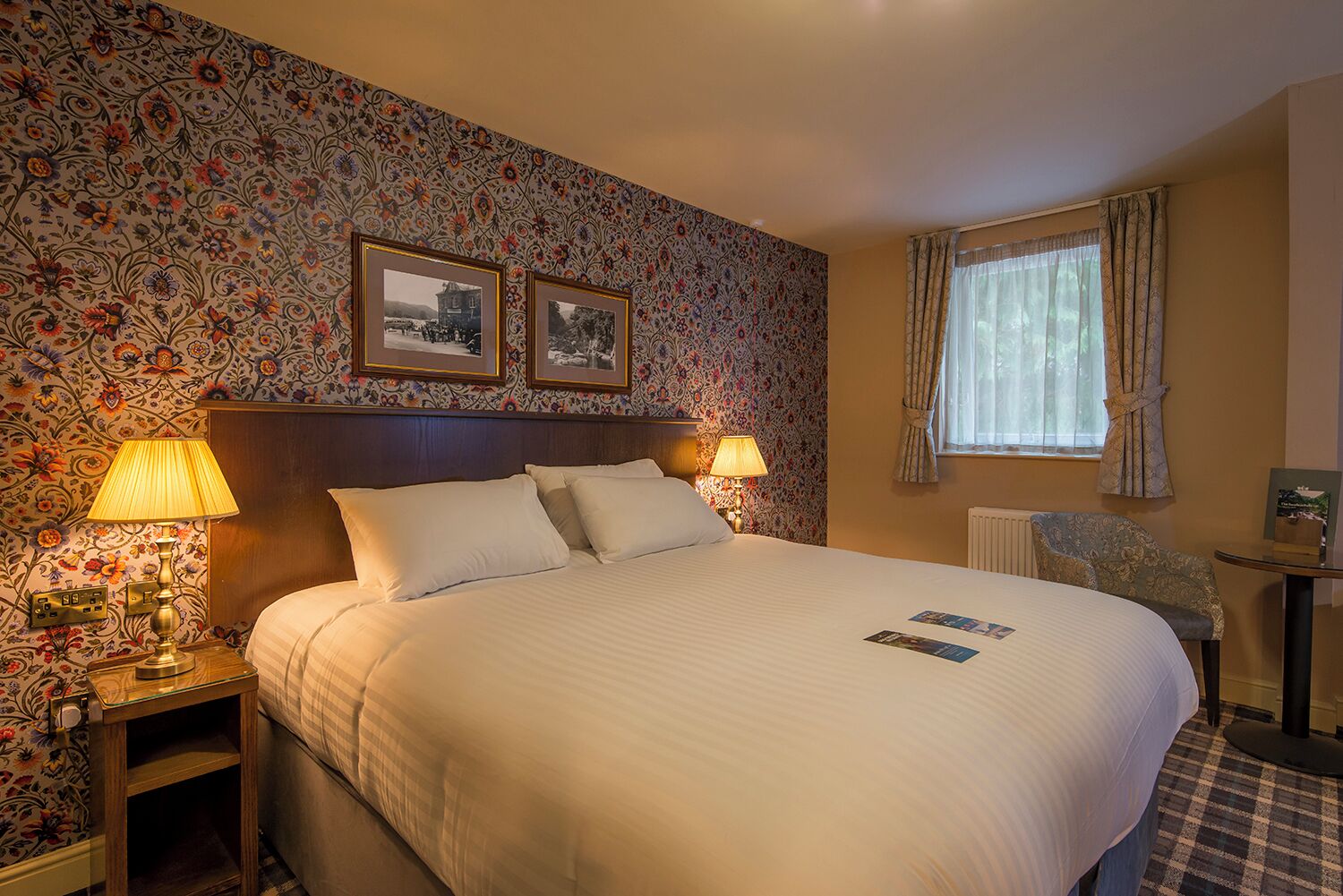 Habitación de hotel con una cama grande, dos lámparas de noche, cabecera de madera, papel tapiz floral, dos cuadros en la pared, una ventana con cortinas y una pequeña mesa con una silla.