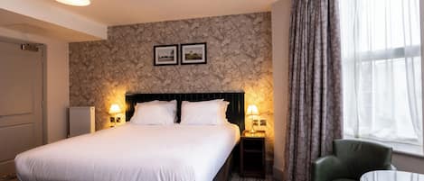 A hotel room with a large bed, two bedside lamps, a small table, a chair, and a window with curtains.