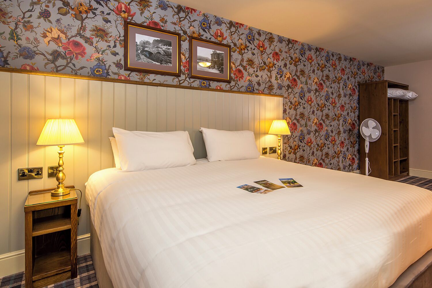 A hotel room with a large bed, two bedside lamps, a wooden nightstand, a fan, and floral wallpaper.