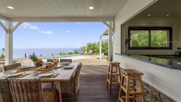 A covered outdoor dining area with a view of the ocean, a wooden table set with plates and cutlery, wicker chairs, and a stone wall. - Villa Kimloa - Three Bedroom Villa, Sleeps 6 (Case-Pilote)