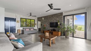 A modern living room with a grey sofa, wooden coffee table, and a stone accent wall. There are ceiling fans, a flat-screen TV, and a large window with a view of the outdoors. - Villa Kimloa - Three Bedroom Villa, Sleeps 6 (Case-Pilote)