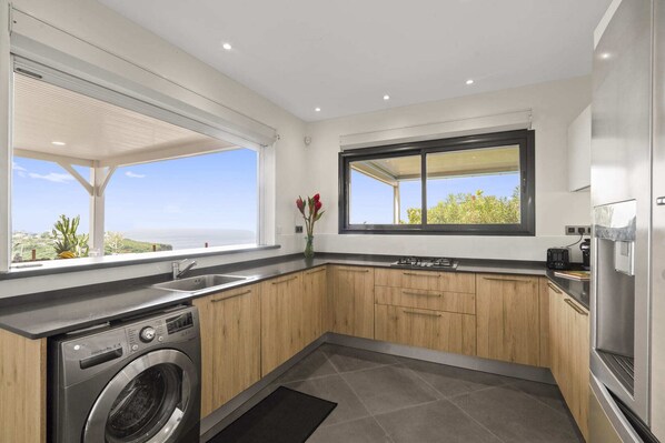 A modern kitchen with wooden cabinets, a built-in washing machine, a large window with a view, and a small sink.