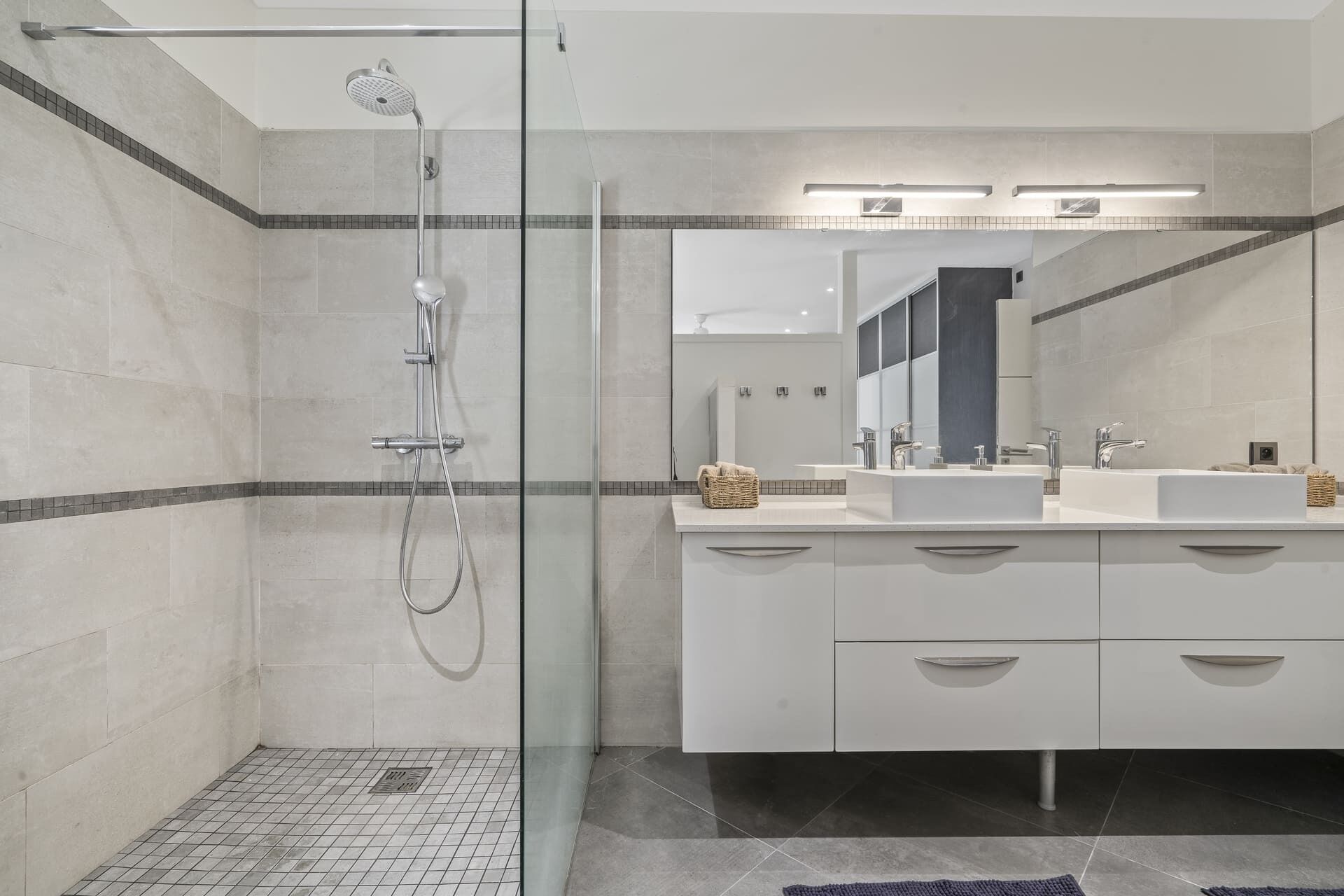Un baño moderno con ducha de vidrio, dos lavamanos y un espejo grande.
