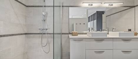 Un baño moderno con ducha de vidrio, dos lavamanos y un espejo grande.