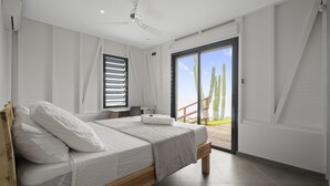 A bedroom with a bed, a ceiling fan, a window with blinds, and a view of cacti through a glass door. - Villa Kimloa - Three Bedroom Villa, Sleeps 6 (Case-Pilote)