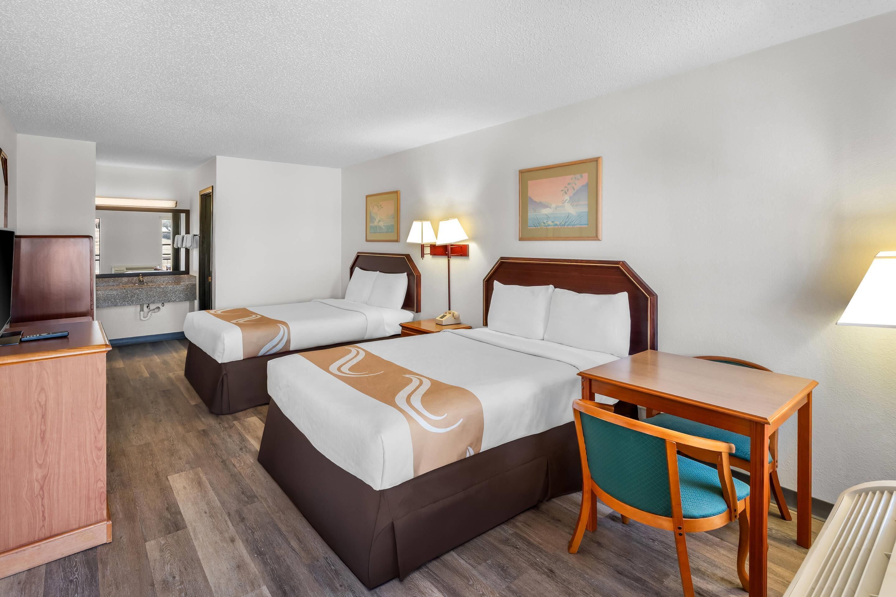 A hotel room with two beds, a desk, a chair, a lamp, and a painting on the wall.