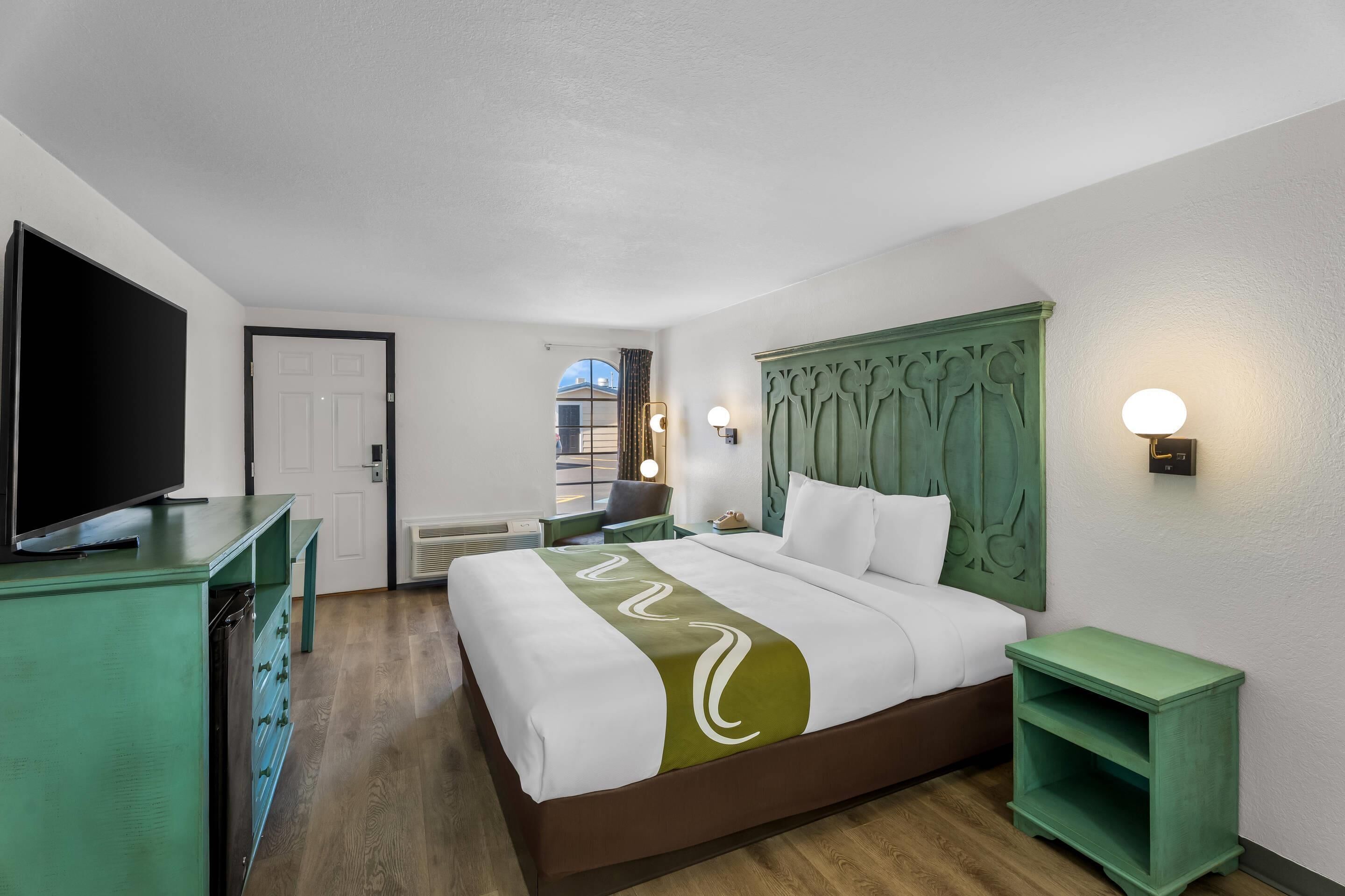 Una habitación de hotel con una cama, un televisor sobre un aparador verde, una mesita de noche con una lámpara y vistas al exterior por una ventana.